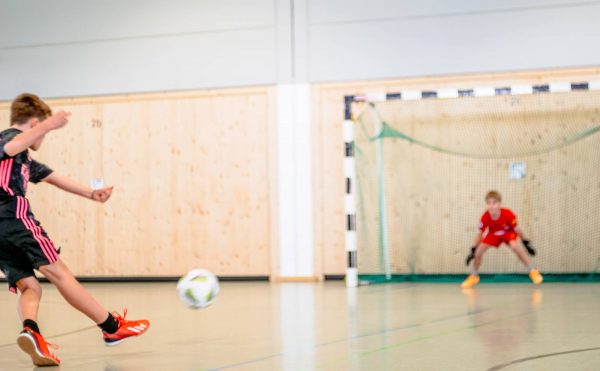 Ein Fußballspieler schießt den Ball auf ein Tor mit Towart in einer Turnhalle.